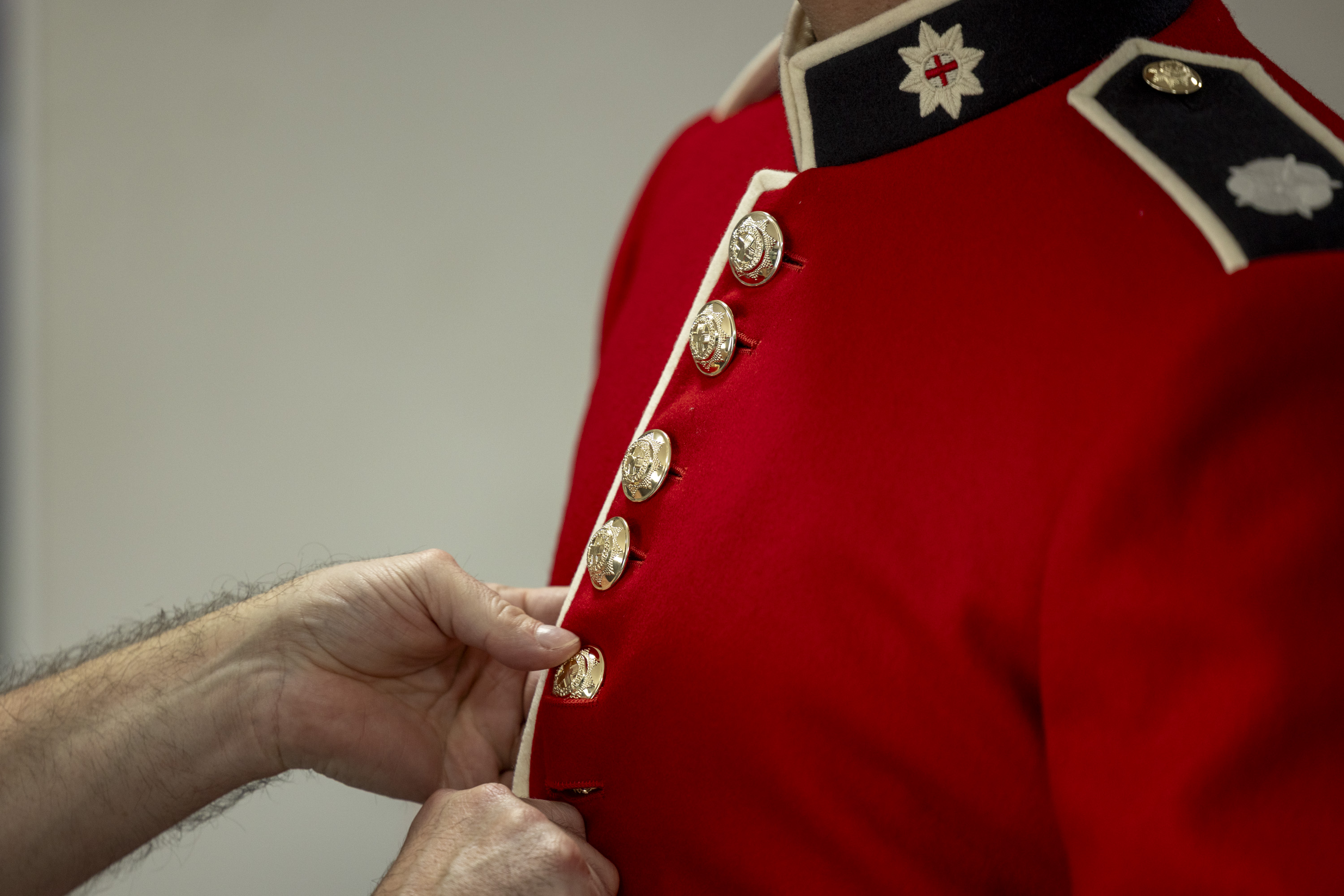 This image shows someone buttoning a red tunic as the ceremonial dress of the Household Division. The jacket features a stand-up collar with white trim and coldstream guards cap badge, and shiny, metallic buttons going down the front of the tunic.