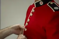 This image shows someone buttoning a red tunic as the ceremonial dress of the Household Division. The jacket features a stand-up collar with white trim and coldstream guards cap badge, and shiny, metallic buttons going down the front of the tunic.