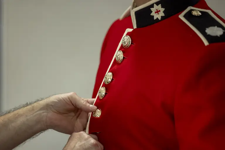 This image shows someone buttoning a red tunic as the ceremonial dress of the Household Division. The jacket features a stand-up collar with white trim and coldstream guards cap badge, and shiny, metallic buttons going down the front of the tunic.