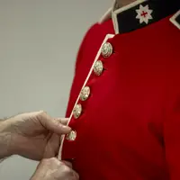 This image shows someone buttoning a red tunic as the ceremonial dress of the Household Division. The jacket features a stand-up collar with white trim and coldstream guards cap badge, and shiny, metallic buttons going down the front of the tunic.