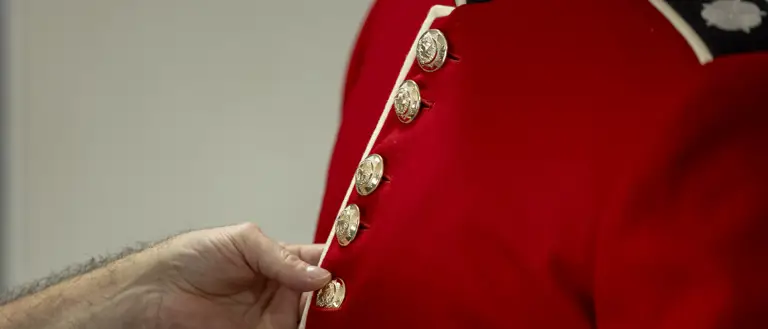 This image shows someone buttoning a red tunic as the ceremonial dress of the Household Division. The jacket features a stand-up collar with white trim and coldstream guards cap badge, and shiny, metallic buttons going down the front of the tunic.