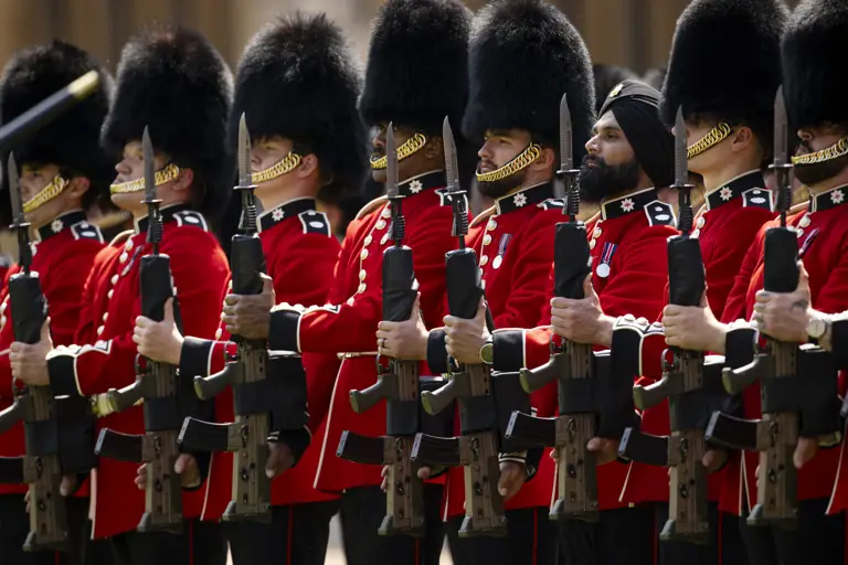 British guards in traditional red uniforms and bearskin hats standing in formation holding rifles.