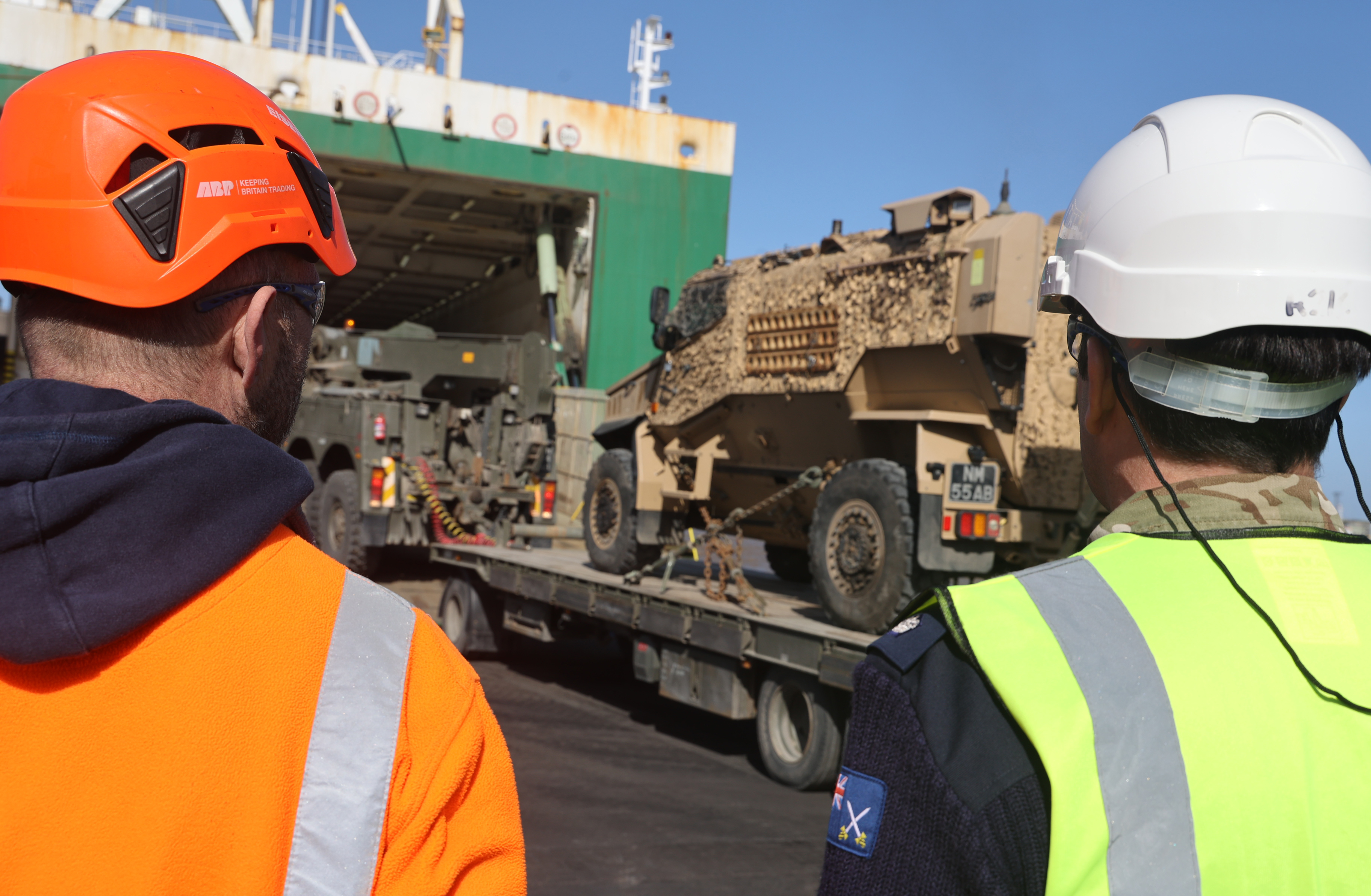 Two men in white hard hats and high vis jackets watch an Army vehicle being loaded onto a ship.