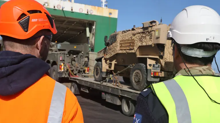 Two men in white hard hats and high vis jackets watch an Army vehicle being loaded onto a ship.