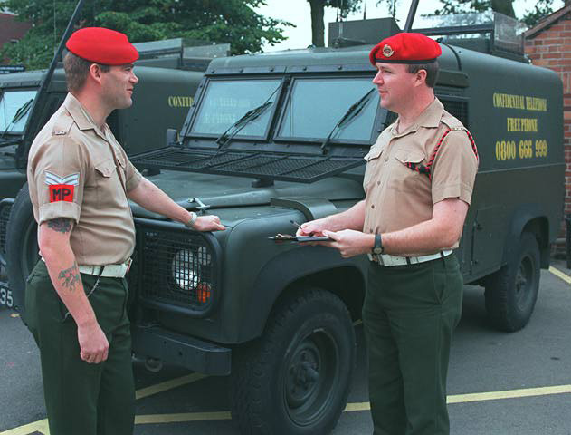 Two individuals in military attire stand in front of army vehicles, both wearing red berets. One holds a clipboard. The vehicles are marked with 