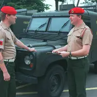 Two individuals in military attire stand in front of army vehicles, both wearing red berets. One holds a clipboard. The vehicles are marked with