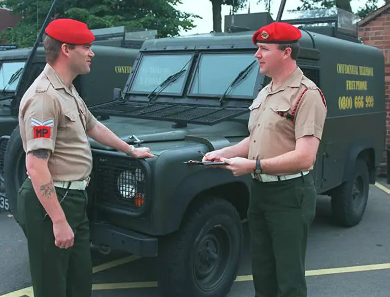 Two individuals in military attire stand in front of army vehicles, both wearing red berets. One holds a clipboard. The vehicles are marked with