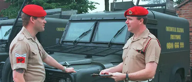 Two individuals in military attire stand in front of army vehicles, both wearing red berets. One holds a clipboard. The vehicles are marked with