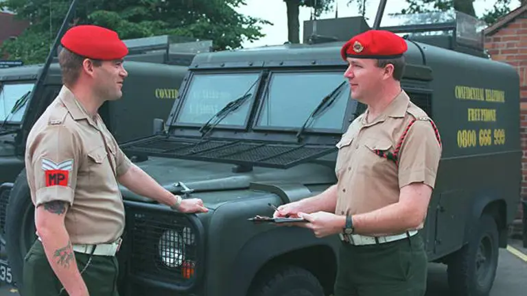 Two individuals in military attire stand in front of army vehicles, both wearing red berets. One holds a clipboard. The vehicles are marked with