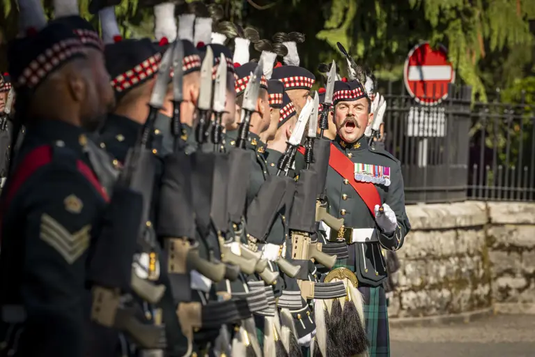 A line of soldiers in traditional Scottish military uniforms hold rifles with fixed bayonets during a formal ceremony.
