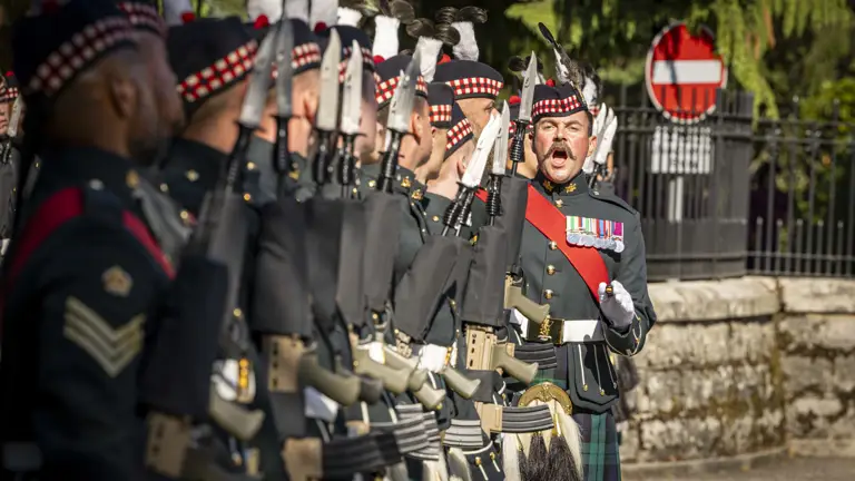 A line of soldiers in traditional Scottish military uniforms hold rifles with fixed bayonets during a formal ceremony.