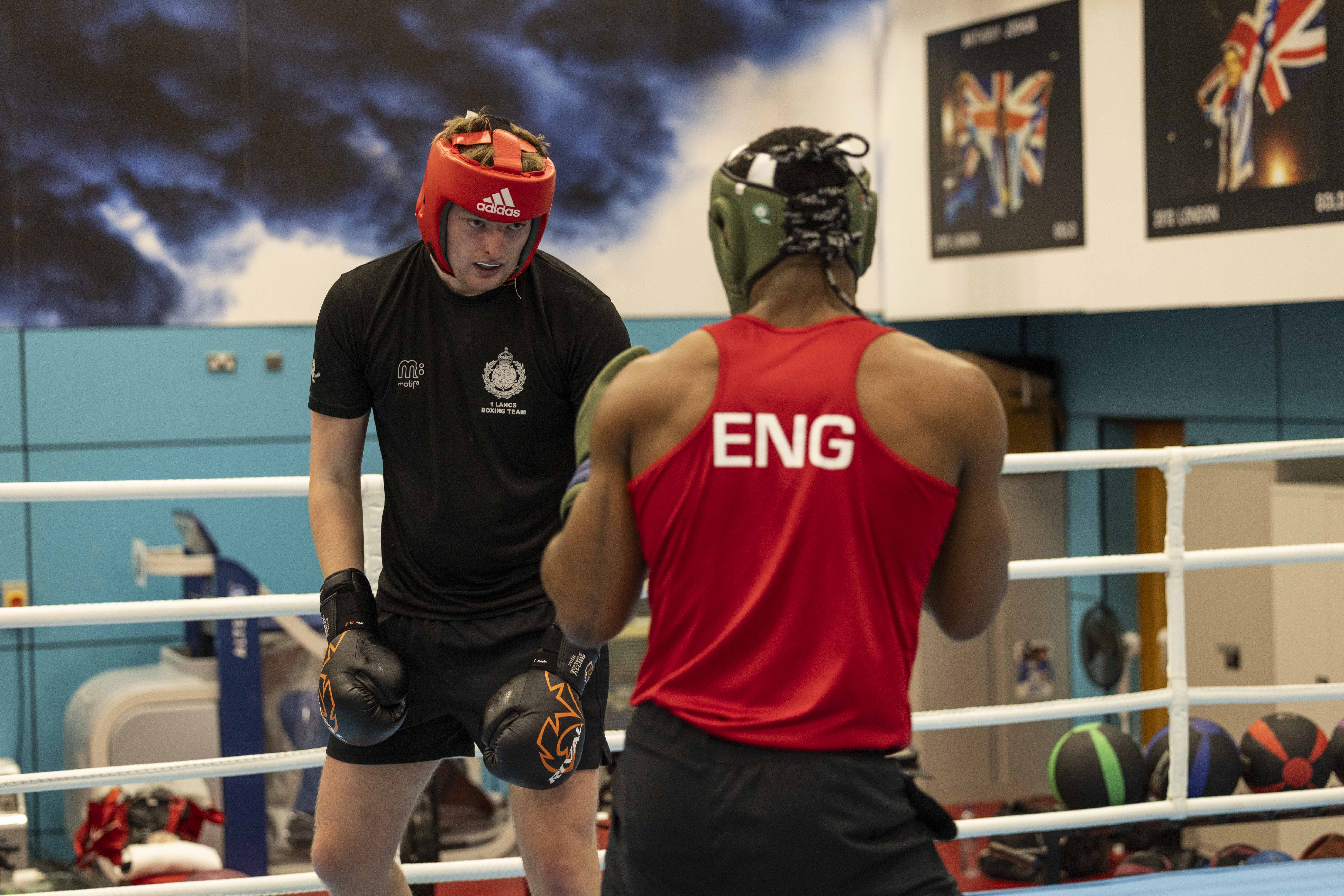 Two men are boxing in a ring. The furthest away wears a black t-shirt and red hat, the closest wears a red top with 'Eng' written on the back, and a green hat.