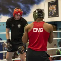 Two men are boxing in a ring. The furthest away wears a black t-shirt and red hat, the closest wears a red top with 'Eng' written on the back, and a green hat.