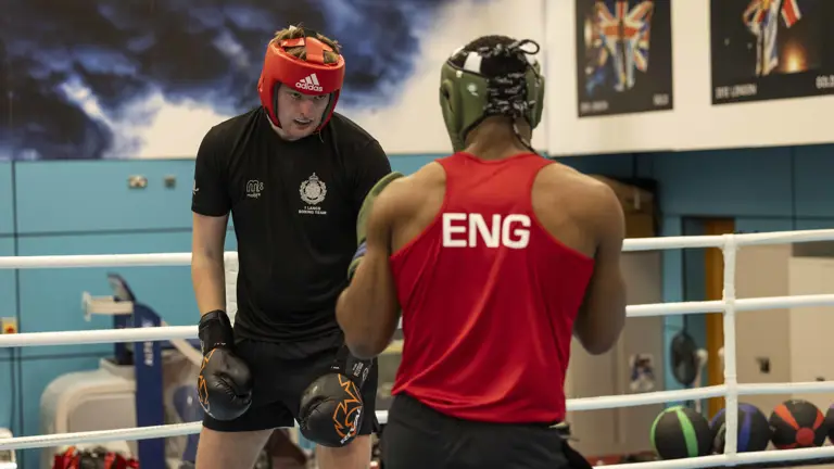 Two men are boxing in a ring. The furthest away wears a black t-shirt and red hat, the closest wears a red top with 'Eng' written on the back, and a green hat.