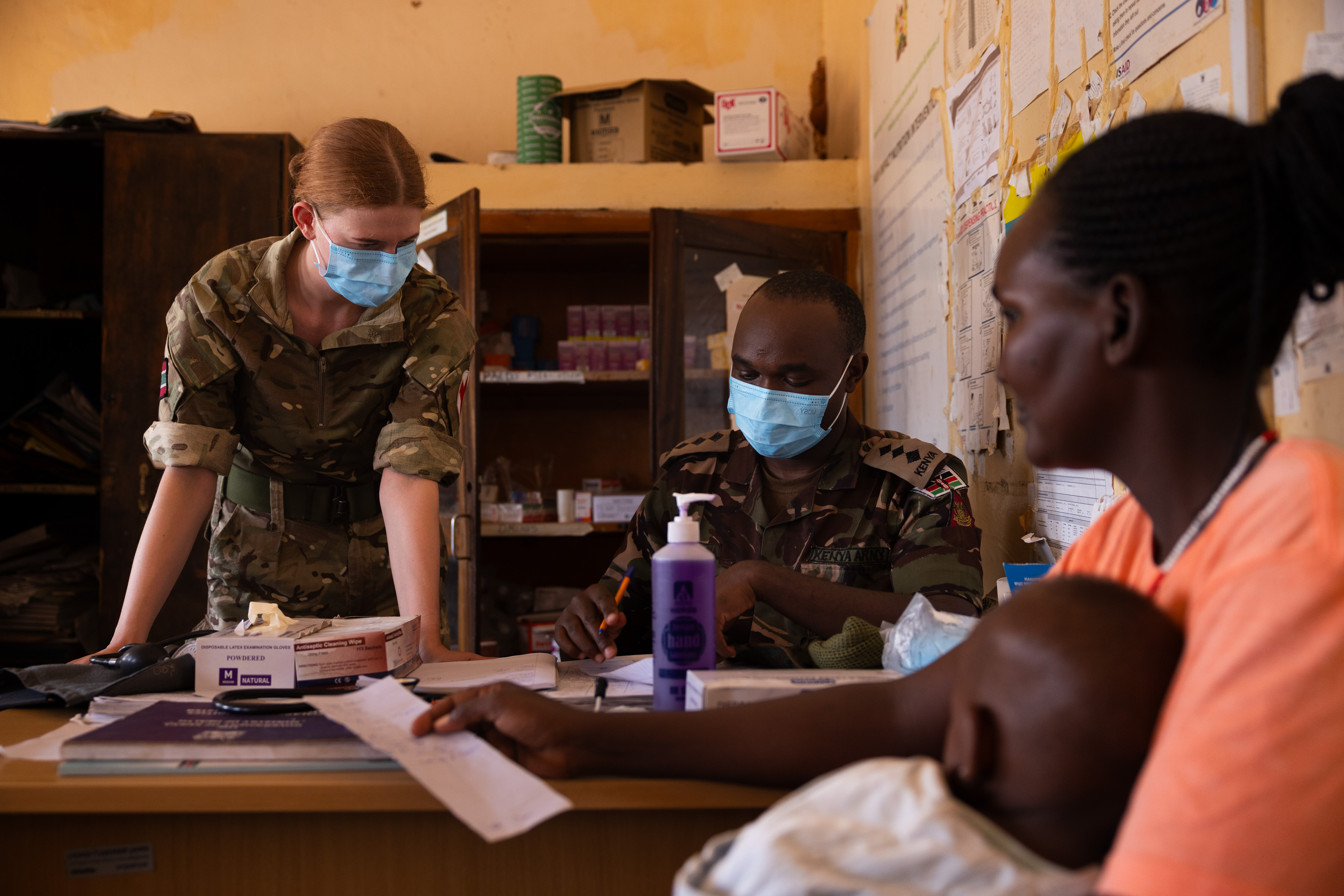 Two uniformed personnel assist a woman holding a baby in a modest office with medical supplies.