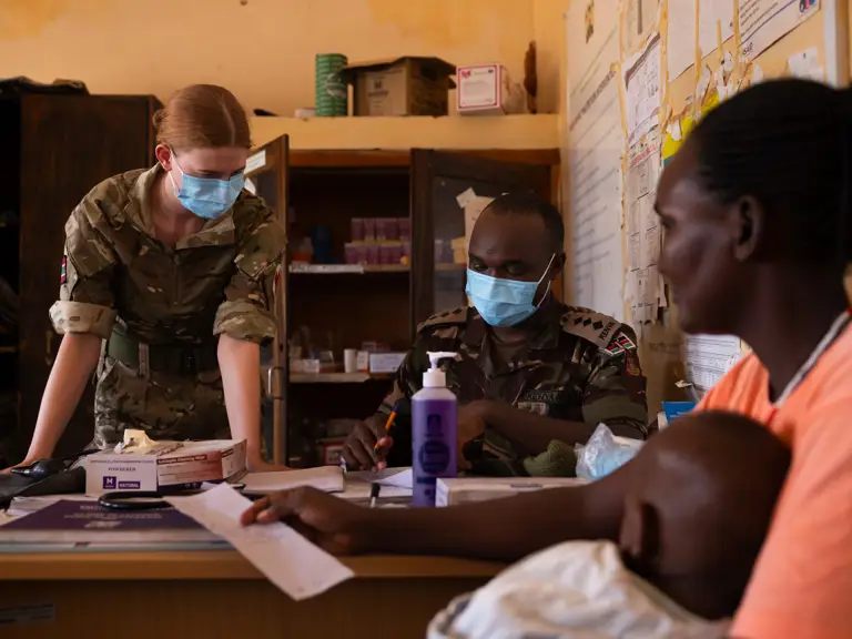 Two uniformed personnel assist a woman holding a baby in a modest office with medical supplies.