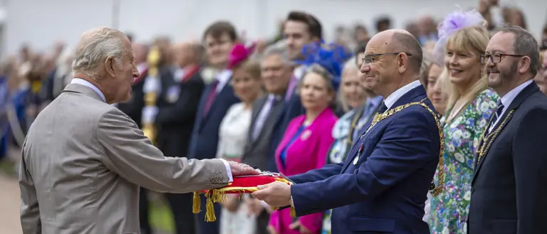 His Majesty The King places his hand onto a red pillow as a crowd looks on.