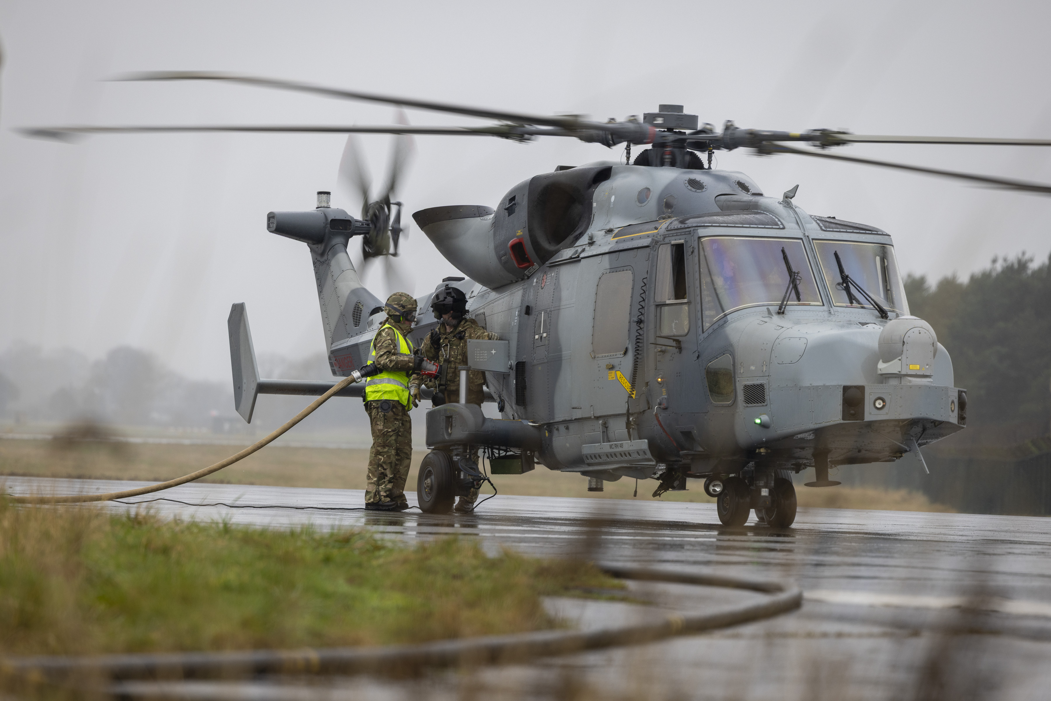 Military personnel refuel a Wildcat helicopter on a foggy airfield. The helicopter is on the ground.