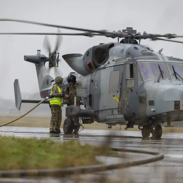 Military personnel refuel a Wildcat helicopter on a foggy airfield. The helicopter is on the ground.