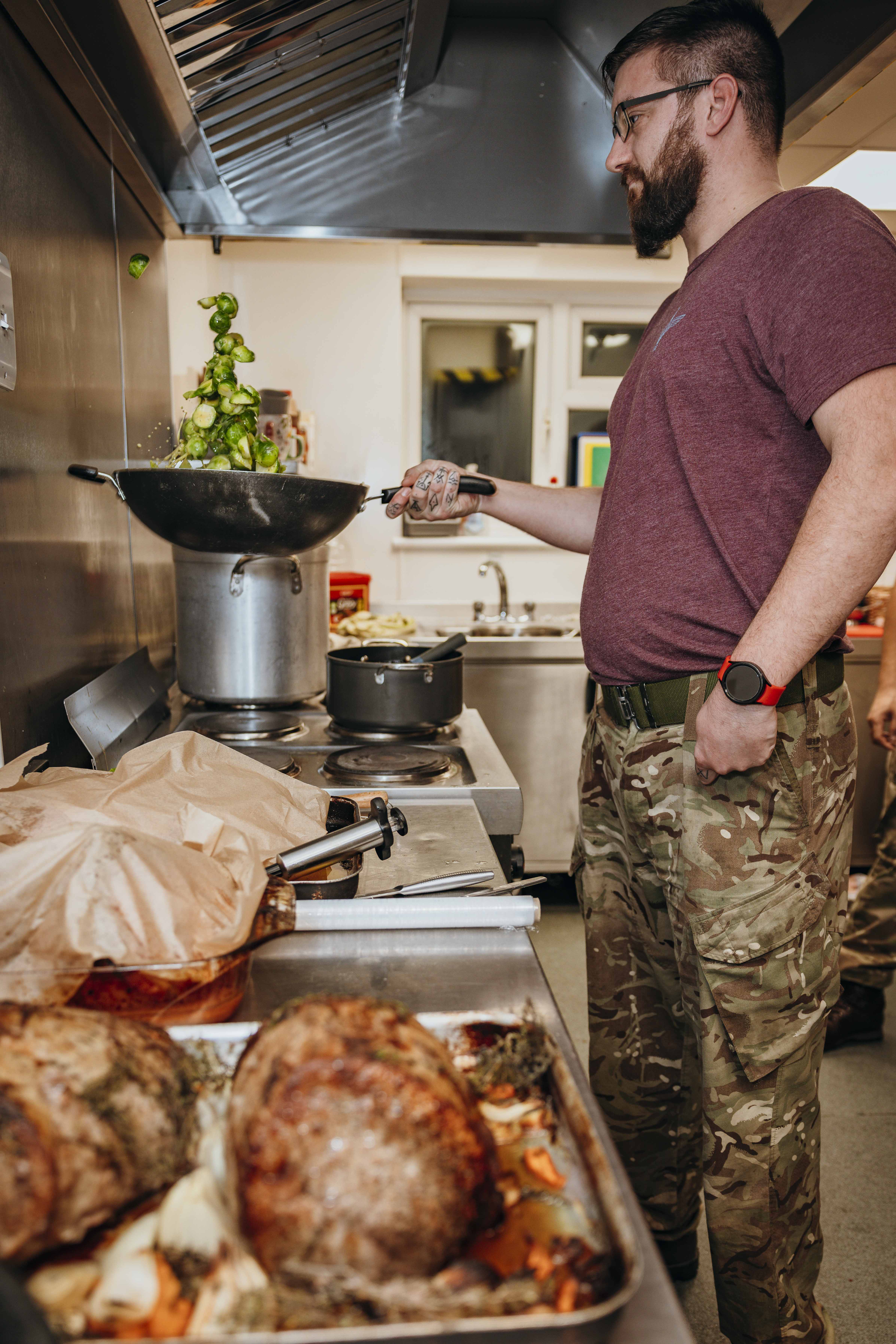 A man in a purple t-shirt and camo trousers prepares brussel sprouts. 