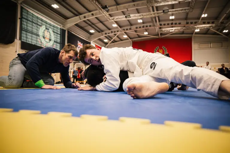 Martial artists practice on a blue mat in a gymnasium. A person in a white gi pins another participant to the mat, while a third individual observes closely. The background features flags and banners.