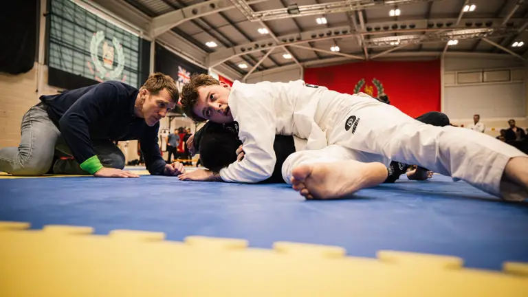 Martial artists practice on a blue mat in a gymnasium. A person in a white gi pins another participant to the mat, while a third individual observes closely. The background features flags and banners.