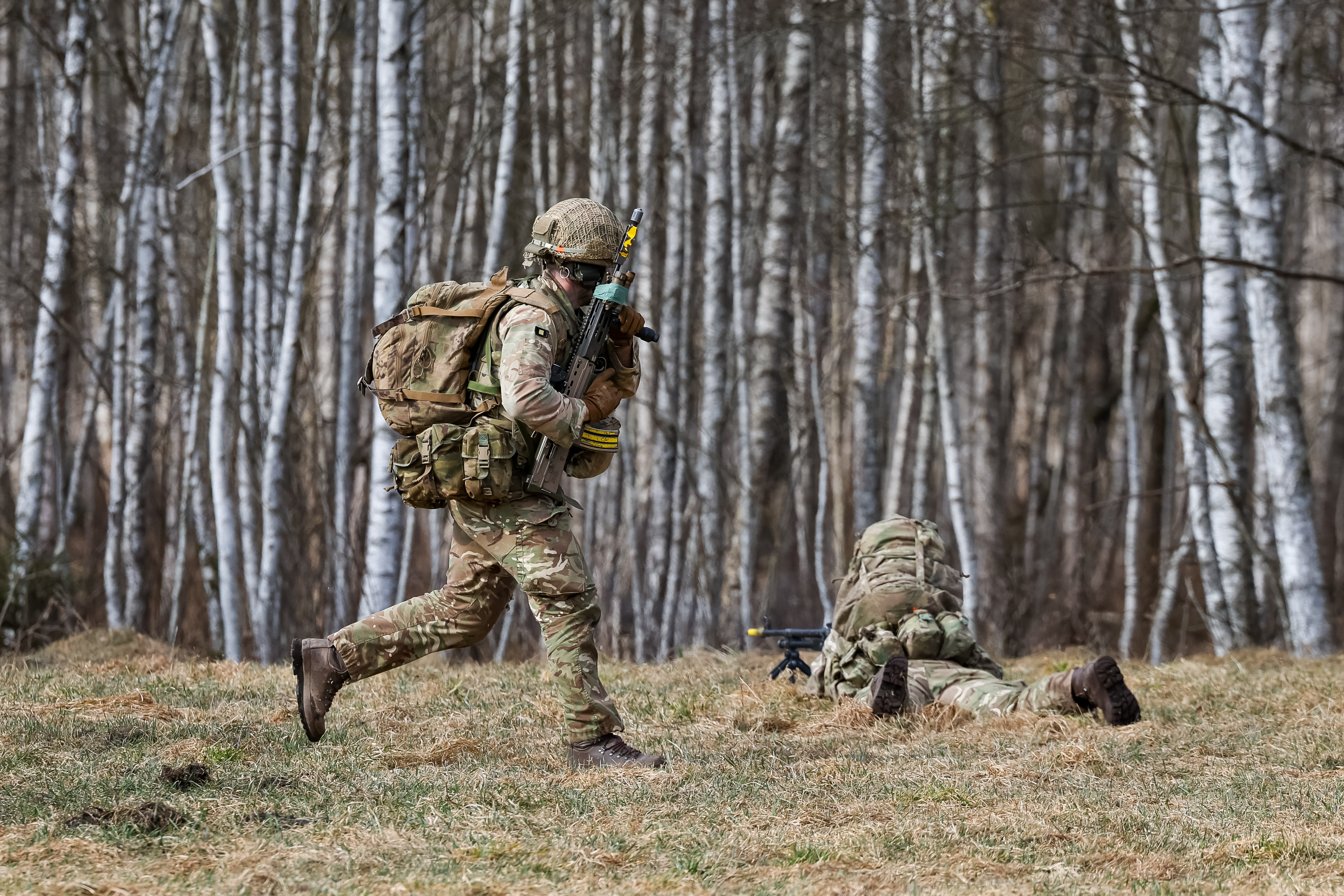 A soldier in camouflage uniform runs while carrying a rifle. On the ground in front of him, another soldier lays down firing a rifle.