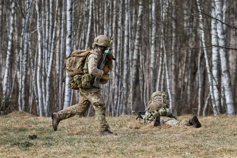 A soldier in camouflage uniform runs while carrying a rifle. On the ground in front of him, another soldier lays down firing a rifle.
