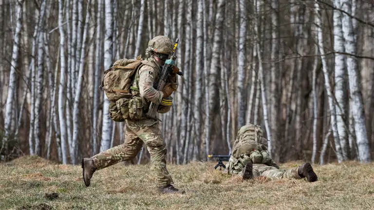 A soldier in camouflage uniform runs while carrying a rifle. On the ground in front of him, another soldier lays down firing a rifle.
