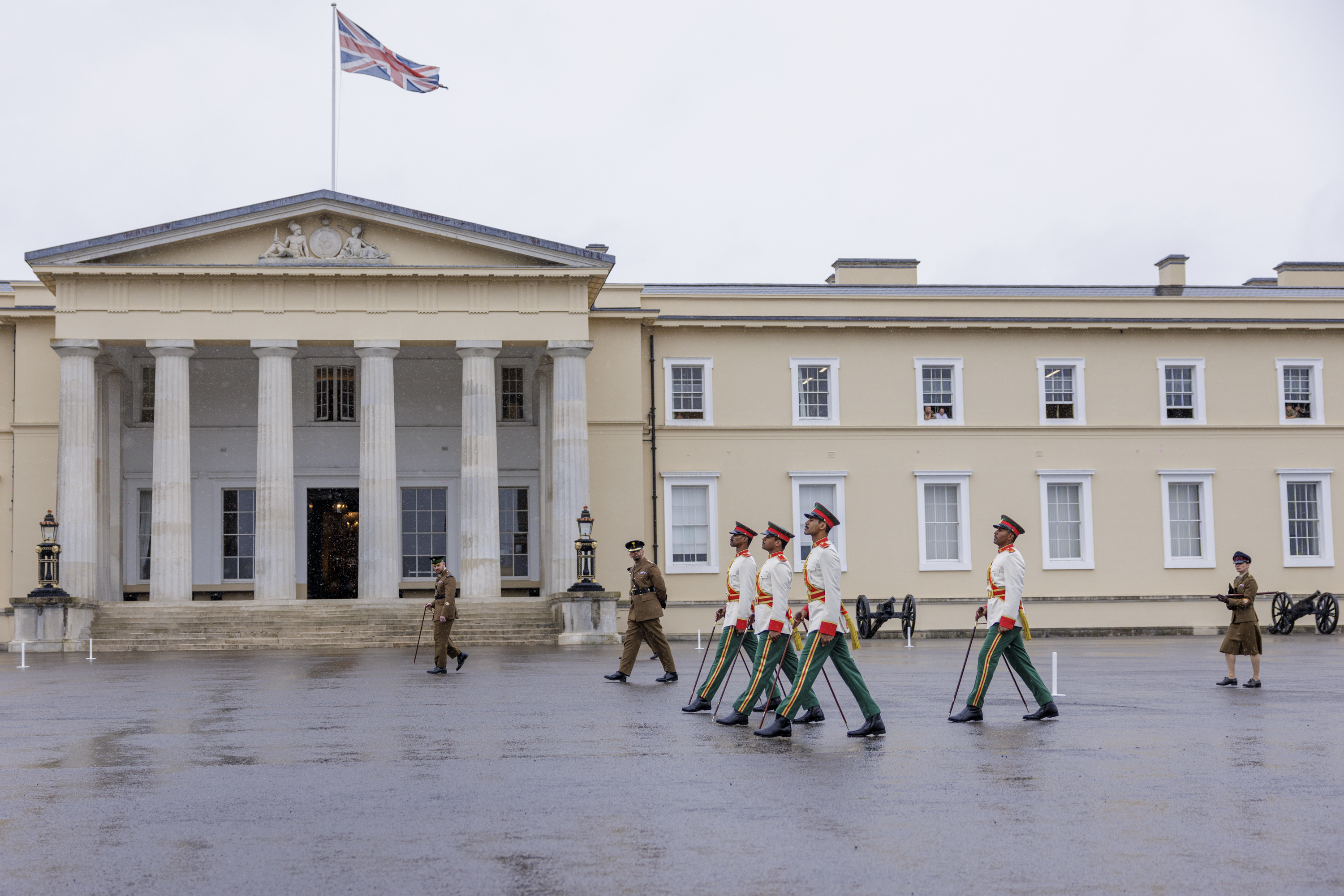 Military personnel in formal uniforms march in formation in front of a large, historic building with columns and the Union Jack flying on top. Additional personnel stand nearby, and there are cannons positioned on the ground.