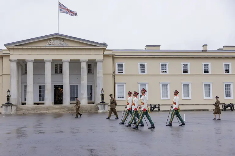 Military personnel in formal uniforms march in formation in front of a large, historic building with columns and the Union Jack flying on top. Additional personnel stand nearby, and there are cannons positioned on the ground.