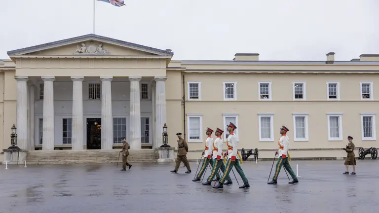 Military personnel in formal uniforms march in formation in front of a large, historic building with columns and the Union Jack flying on top. Additional personnel stand nearby, and there are cannons positioned on the ground.