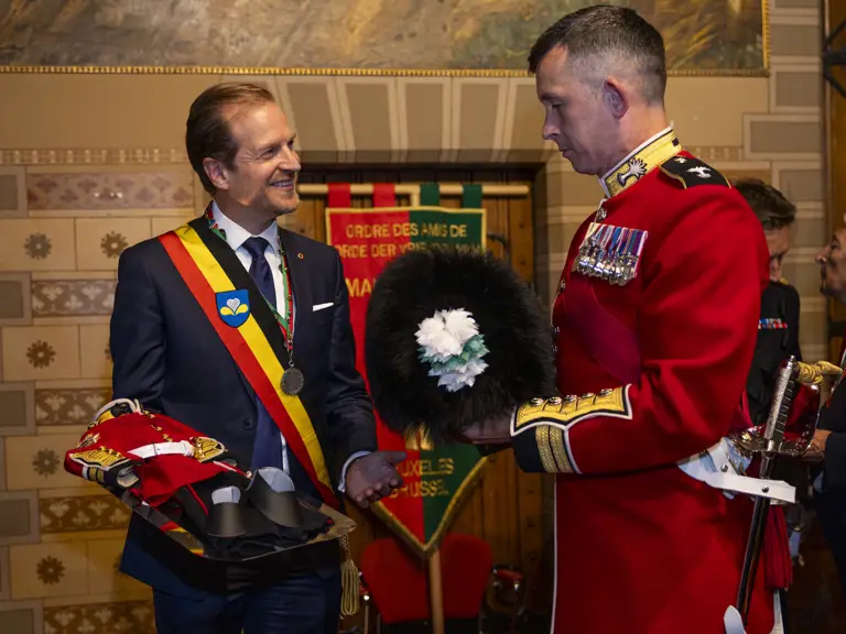 A man in a suit with a red, yellow, and black sash holds a tray with ceremonial military hats, facing a uniformed officer.