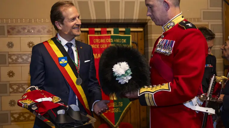 A man in a suit with a red, yellow, and black sash holds a tray with ceremonial military hats, facing a uniformed officer.