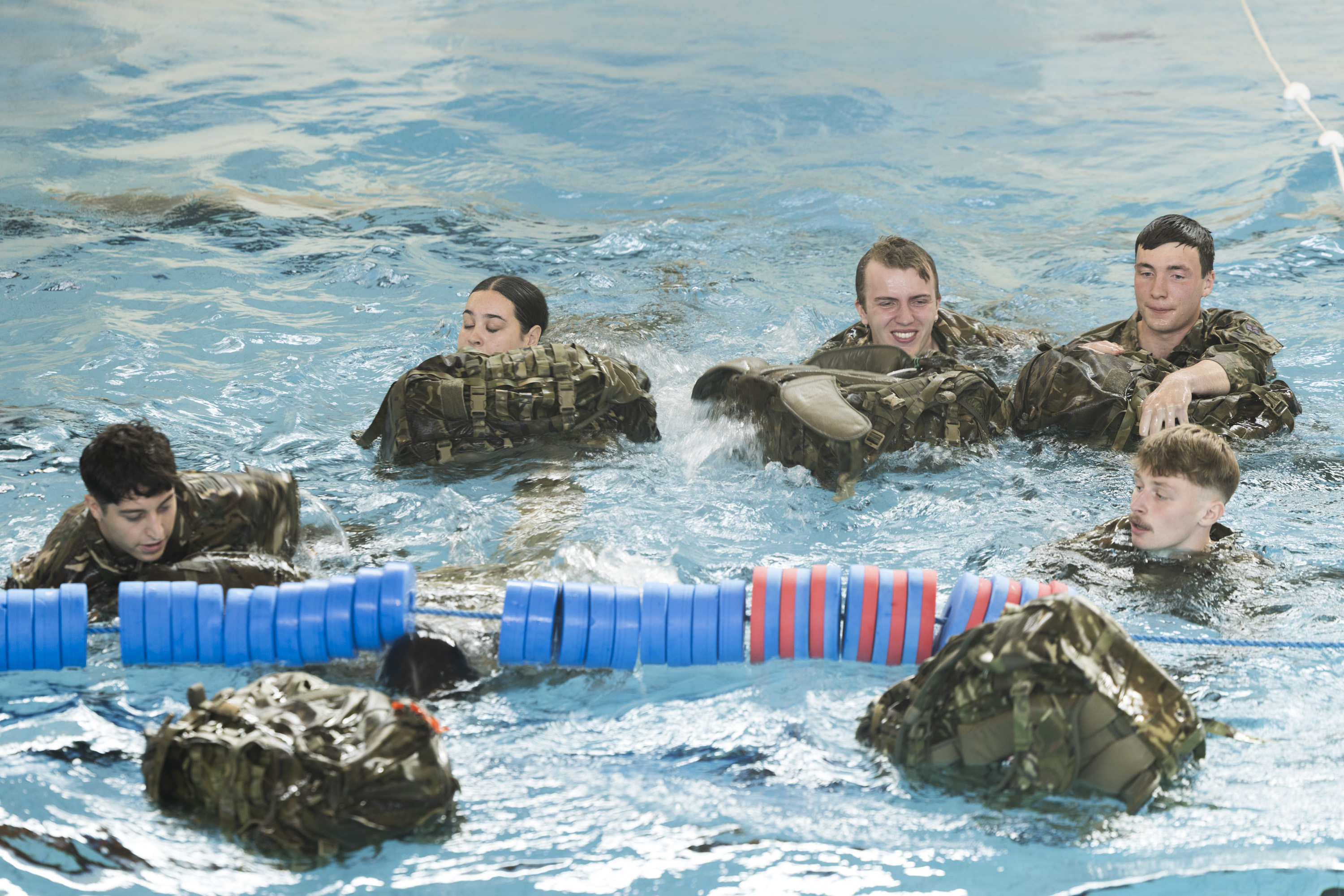 Group of soldiers in camouflage uniforms practicing water survival skills in a pool, partially submerged.