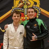 Two men stand side by side, smiling. One wears a white martial arts gi, the other a black and green sports jacket. A lion emblem is in the background.