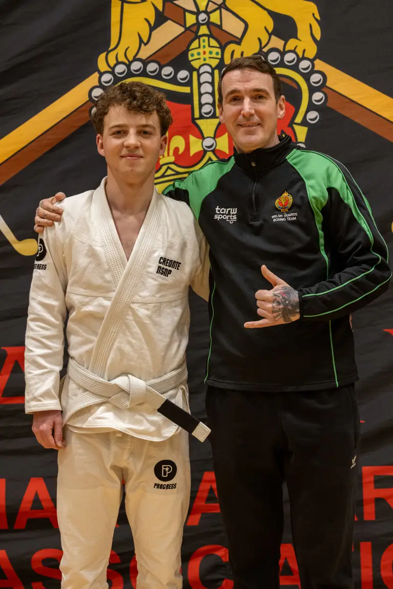 Two men stand side by side, smiling. One wears a white martial arts gi, the other a black and green sports jacket. A lion emblem is in the background.