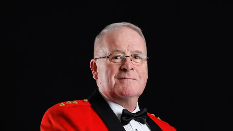 Man in formal military dress uniform with red jacket, black lapels, and medals standing against a black background.