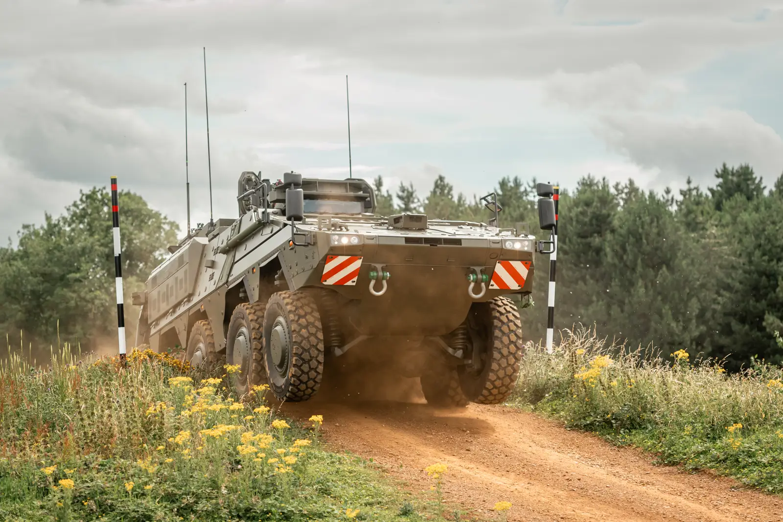 A military armoured vehicle is driving on a dirt road surrounded by grass and yellow wildflowers.