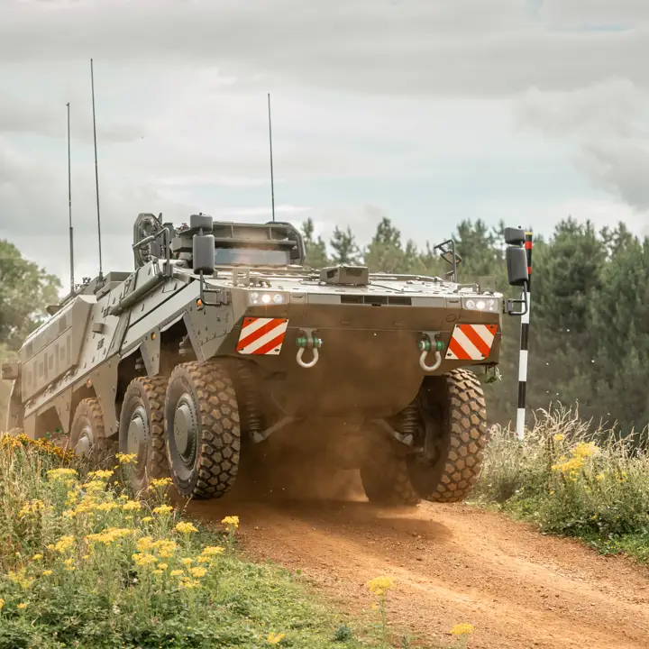 A military armoured vehicle is driving on a dirt road surrounded by grass and yellow wildflowers.