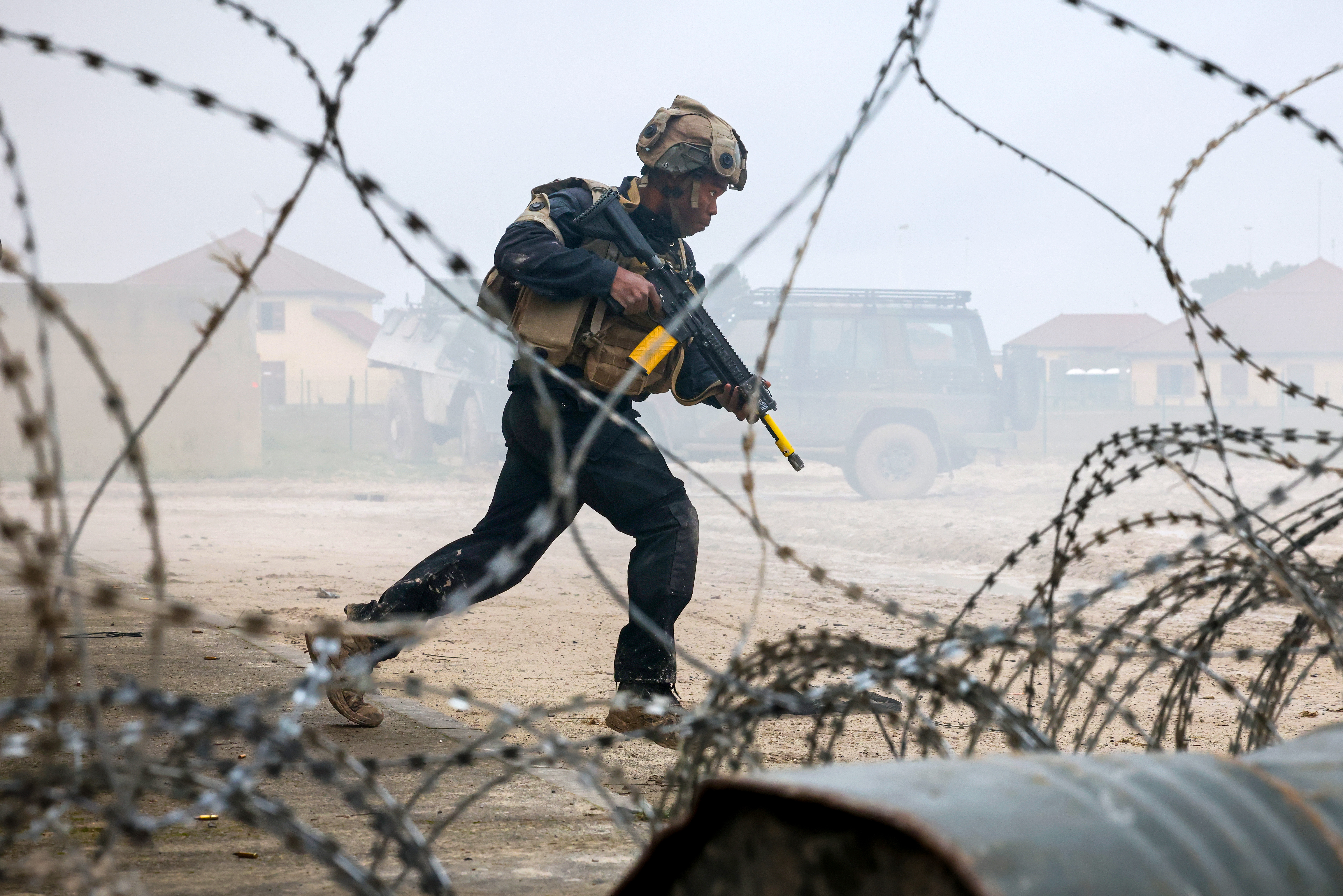 Soldier in tactical gear running with a rifle behind barbed wire in a dusty, foggy environment near military vehicles.