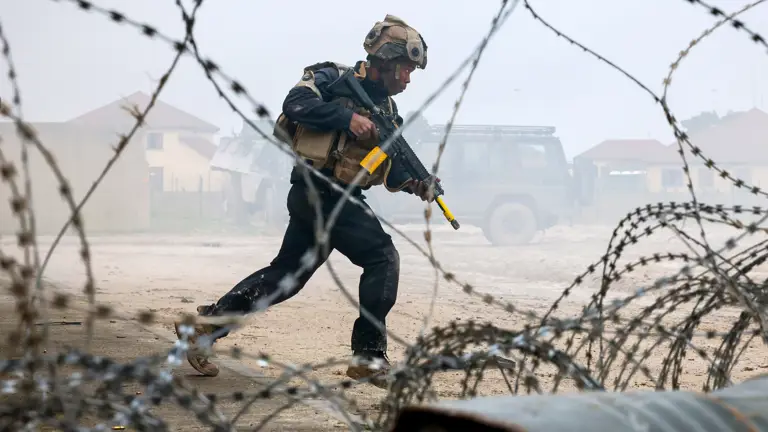 Soldier in tactical gear running with a rifle behind barbed wire in a dusty, foggy environment near military vehicles.