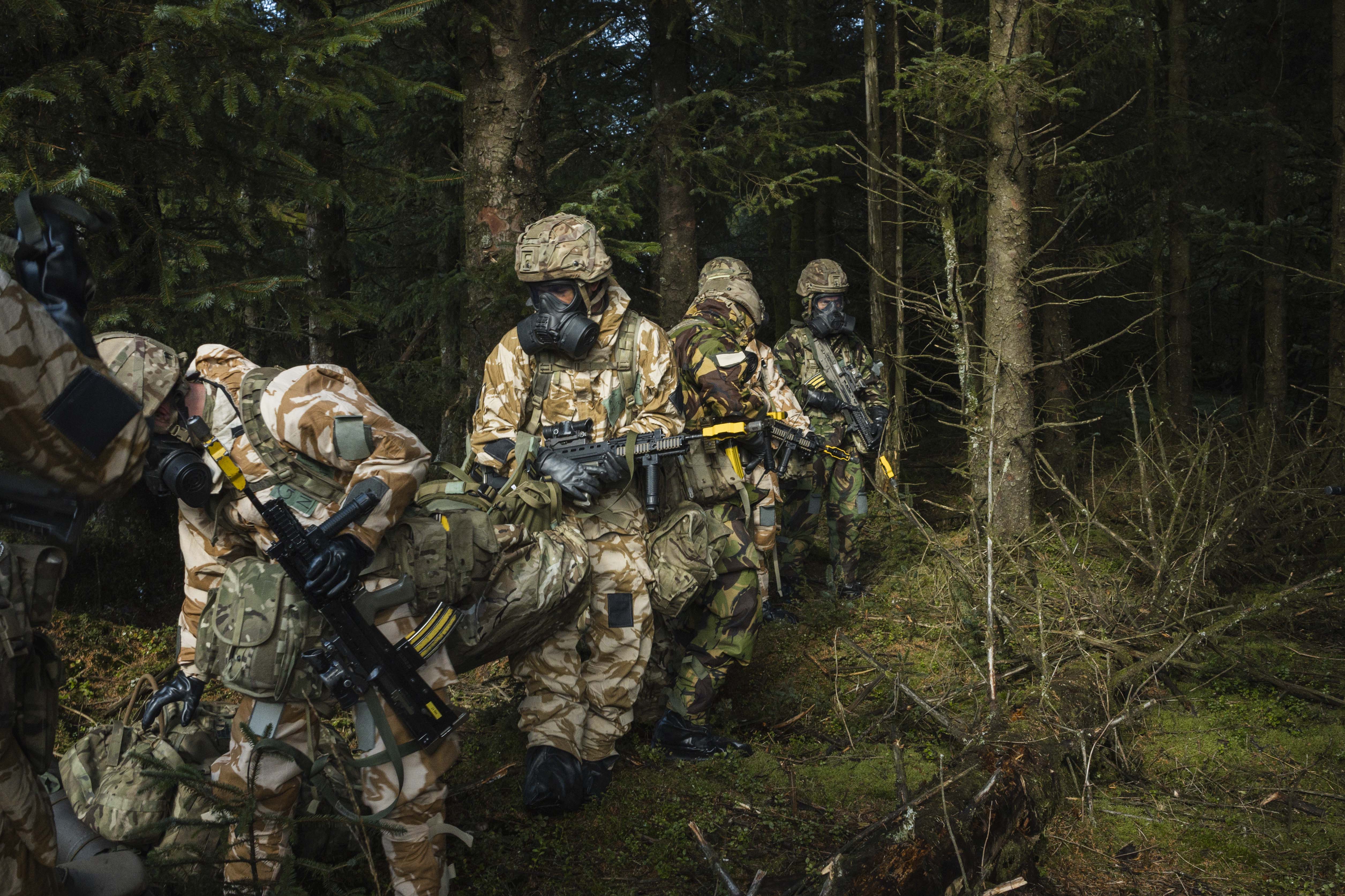 Soldiers in camouflage and gas masks conduct a training exercise in a dense forest. The scene is tense, with weapons ready and focused expressions. 