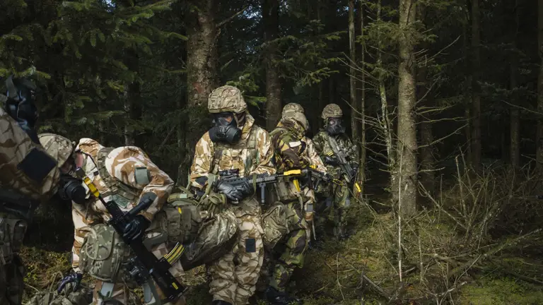 Soldiers in camouflage and gas masks conduct a training exercise in a dense forest. The scene is tense, with weapons ready and focused expressions.
