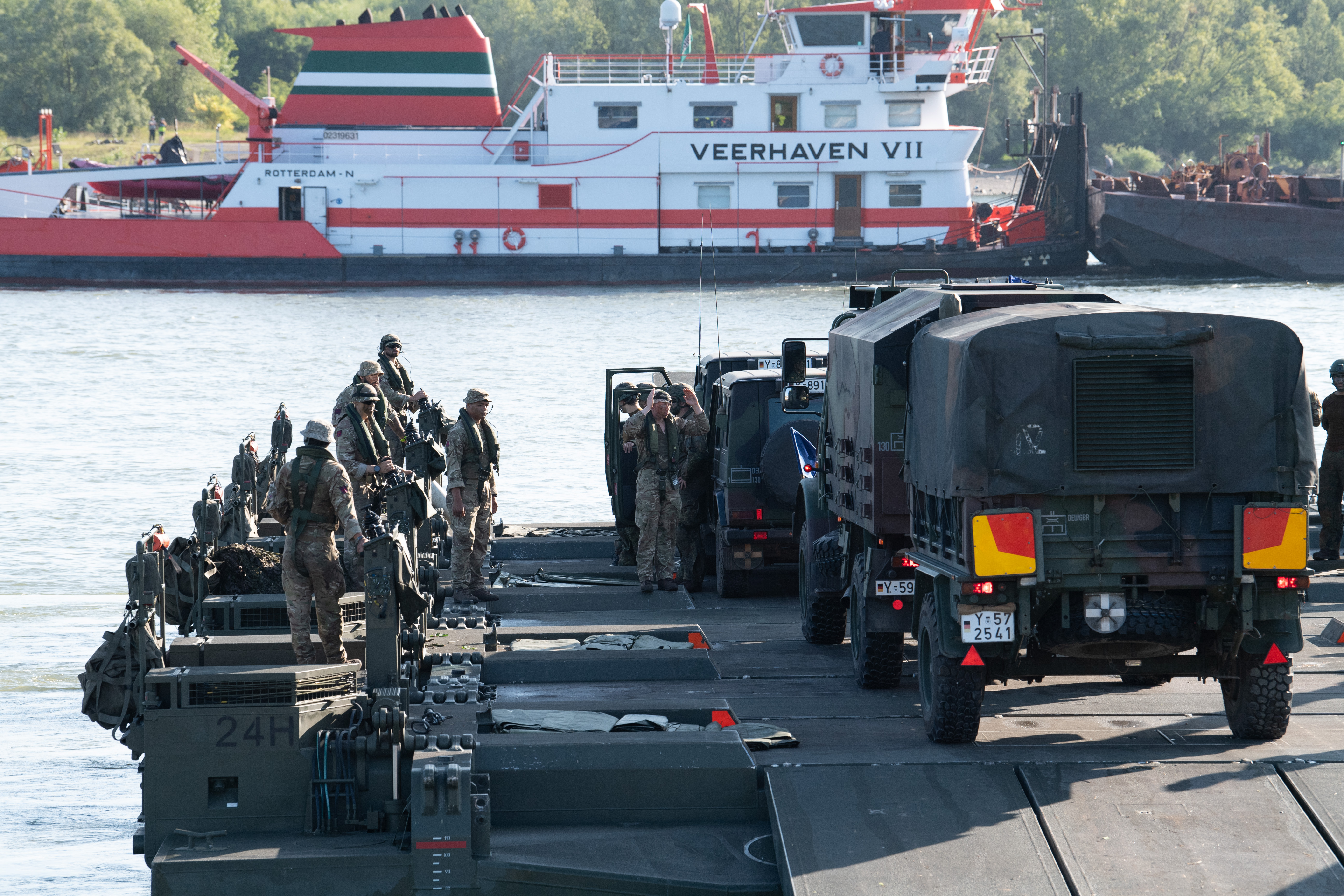 Military personnel stand on a floating bridge as vehicles prepare to cross a river during a training exercise.