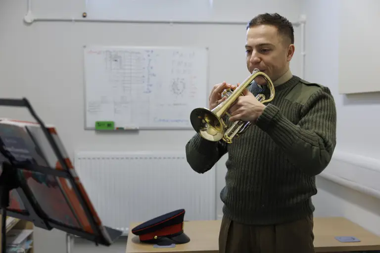 Man in military uniform plays a trumpet in a rehearsal room. A music stand and a peaked cap are visible, with a whiteboard in the background.