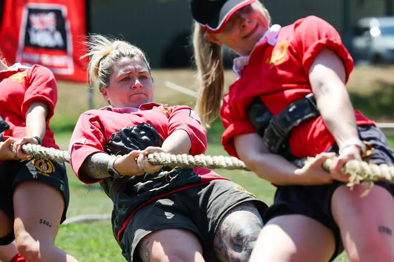 A group of individuals in red shirts participates in a tug-of-war competition. The person in the center is leaning back, pulling the rope with effort. There is a visible tattoo on their leg. A UK Tug of War Association banner is in the background.