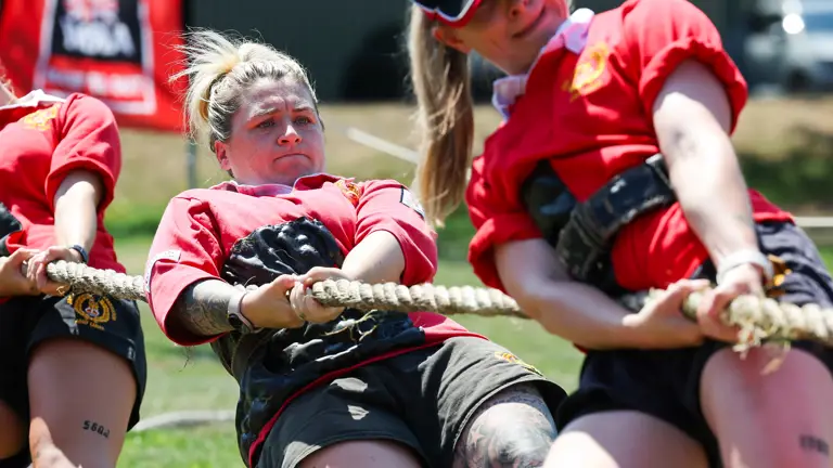 A group of individuals in red shirts participates in a tug-of-war competition. The person in the center is leaning back, pulling the rope with effort. There is a visible tattoo on their leg. A UK Tug of War Association banner is in the background.