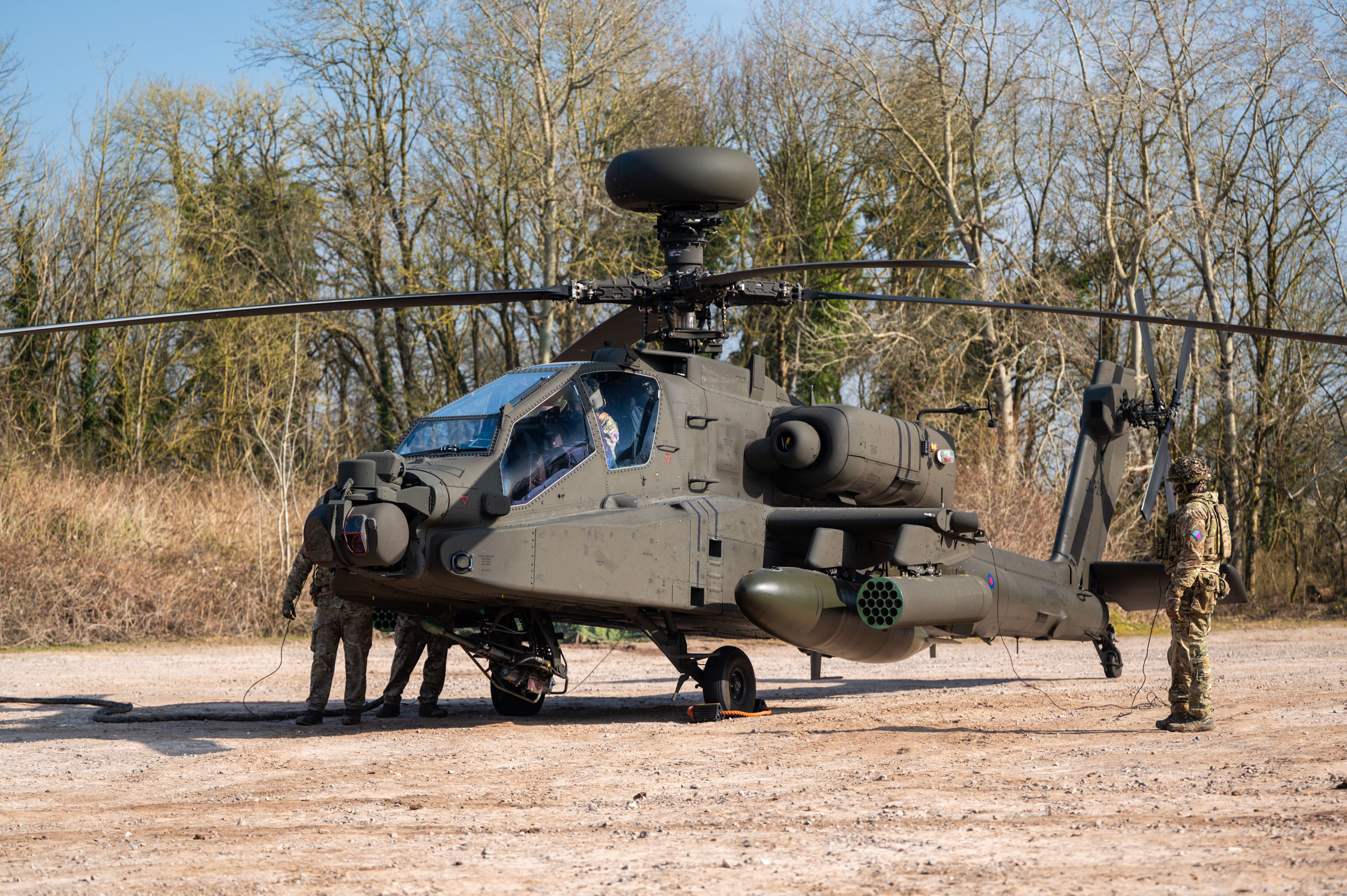 Military personnel inspecting a camouflaged attack helicopter parked on a dirt clearing near trees.