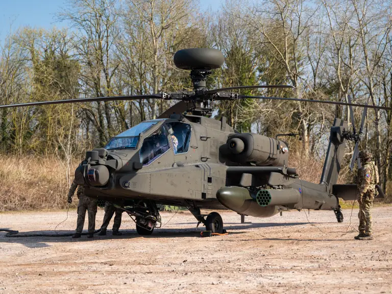 Military personnel inspecting a camouflaged attack helicopter parked on a dirt clearing near trees.
